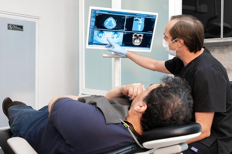 Dr. Sforzina, dentist at SOTA Dental in Cancun, Mexico, pointing to a CT scan x-ray. A patient is laying in the chair and contemplating the dentists decisions.