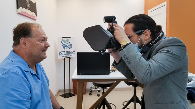 Dr. Nestor, dentist in Los Algodones at SOTA Dental, takes a photo of an older male patient who finished an all-on-4. The patient is smiling, and the doctor is focusing the camera. SOTA Dental sign in the background.