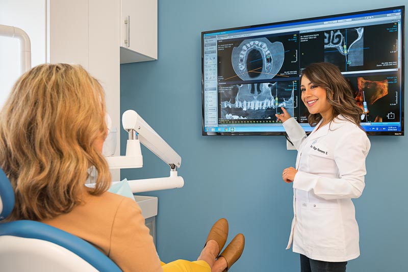 Dr. Olga, dentist in Tijuana at SOTA Dental, speakings with an older female patient. The female patient is sitting in the dental chair and the dentist is pointing to a TV screen with a CT scan.