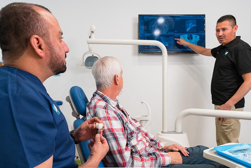 Antonio and Dr. Trejo from SOTA Dental in Tijuana are speaking to an older male patient. Antonio is pointing to a CT scan on a television screen, and the others are listening intently.
