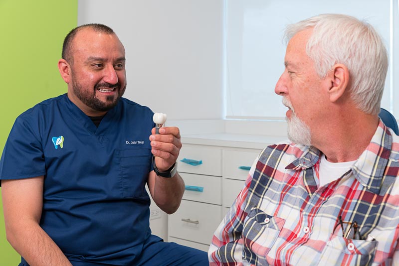Dr. Trejo, dentist in Tijuana at SOTA Dental, is holding a dental implant model while speaking with an older male patient. The doctor is smiling and the patient is speaking to him.