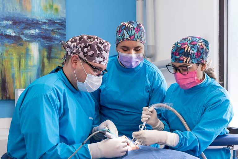 Dr. Bogarin, Dr. Olga, and Dr. Dennise, dentists at SOTA Dental in Tijuana, Mexico, performing surgery. They are all wearing surgical clothes and focusing intently on their work.