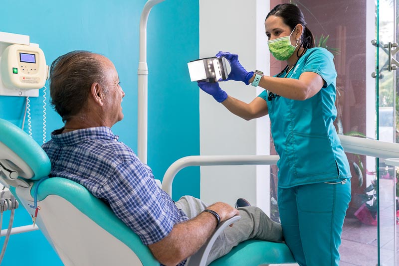 Dr. Alejandra, a dentist at SOTA Dental in Playa del Carmen, takes pictures of a older male patient. She is using a smartphone and camera device and the patient is smiling.