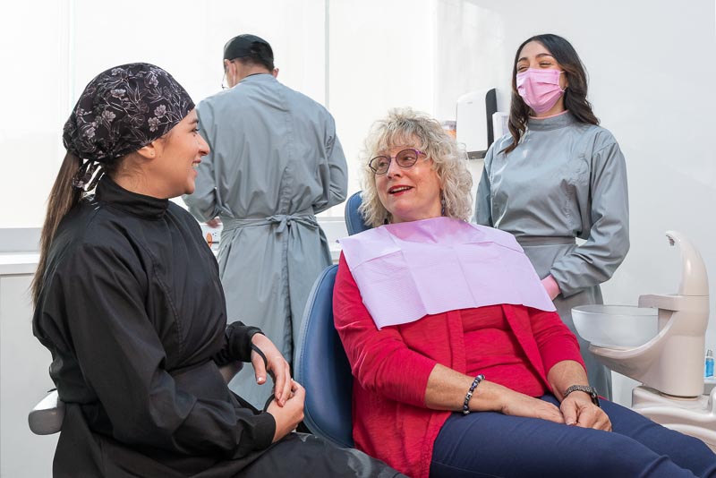 Dr. Alma, dentist in Tijuana at SOTA Dental, speaks with an older female patient. The dentist is laughing as well as the other female dental assistant. One male dental assistant is working in the background.