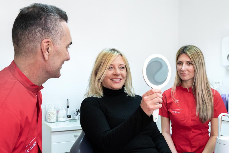 Dr. Jurisic, a dentist for SOTA Dental in Dubrovnik, Croatia, sits with a female patient who is looking her new dental implant in a mirror and smiling.