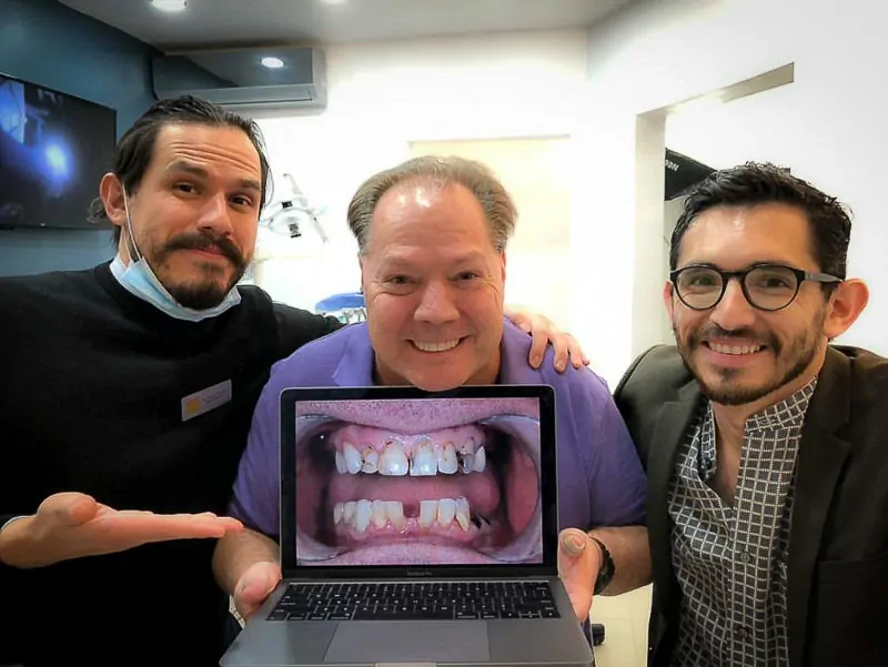 Dr. Nestor and Dr. Diego, dentist in Los Algodones at SOTA Dental Mexico, look at the camera with an older male patient. The patient is holding a laptop computer displaying his teeth before his All on 4 surgery. Everyone is smiling.